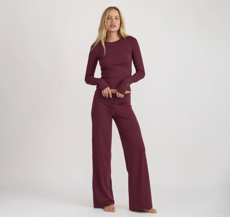 A woman stands barefoot against a plain background, wearing a long-sleeve, burgundy top and matching wide-leg pants.