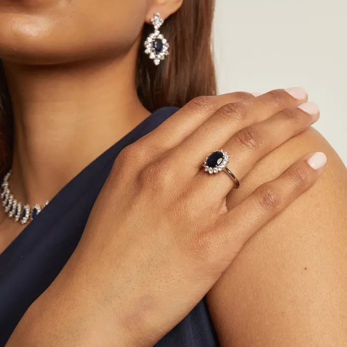 A person wearing a navy dress displays a matching set of jewelry featuring dark blue stones with clear gemstone accents: a necklace, drop earrings, and a ring.
