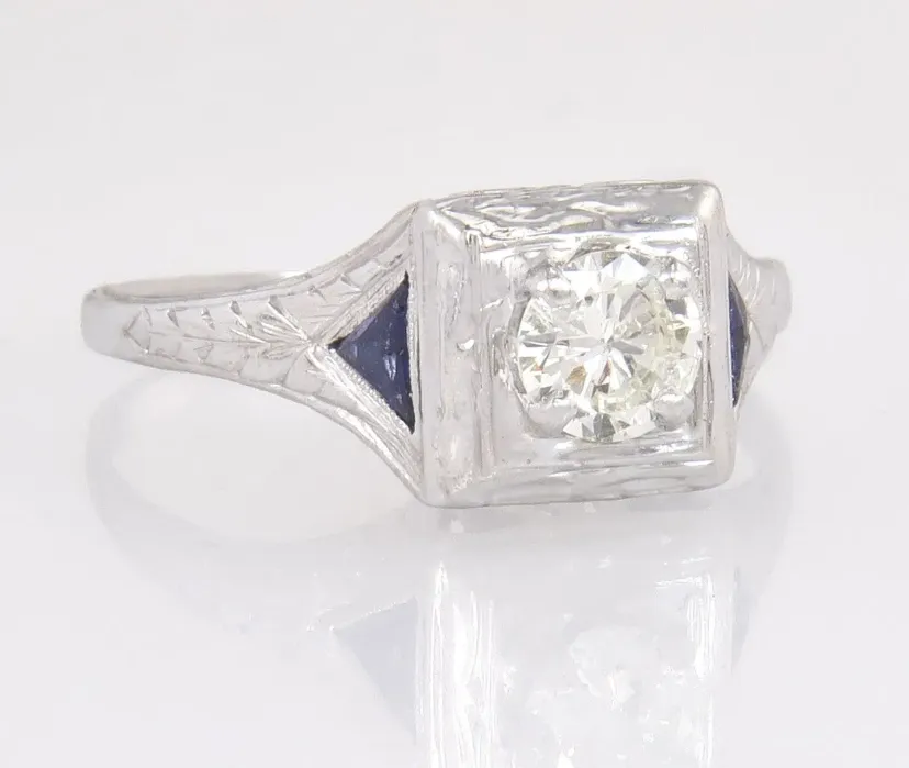 A silver ring with an engraved band features a round, clear gemstone set in a square bezel, flanked by small dark blue triangular accents.