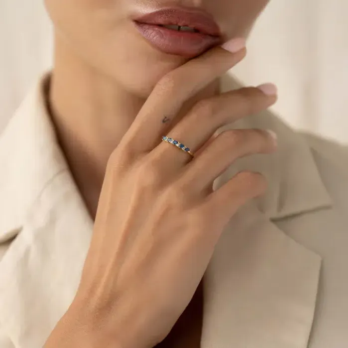 A person wearing a beige blazer touches their face, displaying a gold ring with blue stones on their finger.