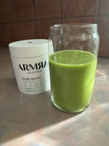 A tub of ARMRA Colostrum powder sits next to a glass filled with a bright green smoothie on a countertop.