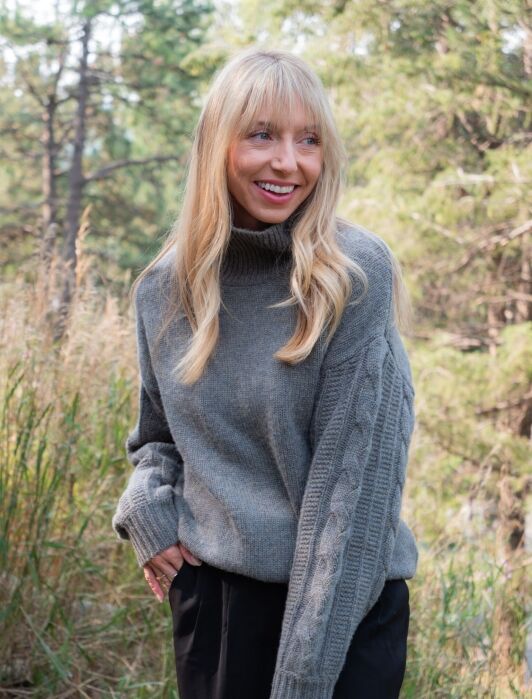 A woman with long blonde hair wearing a gray knit sweater and black pants stands outdoors in a wooded area, smiling.