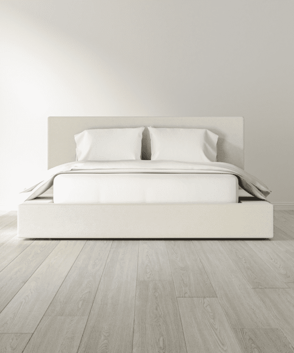 A minimalist bedroom features a beige upholstered bed with white bedding and two pillows, set on light wood flooring against a plain light-colored wall.