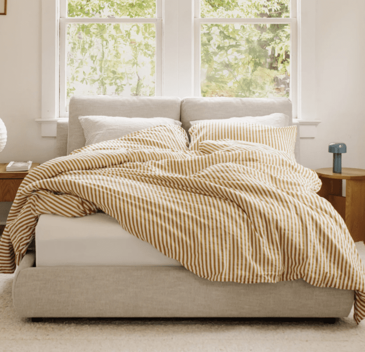 A neatly made bed with yellow and white striped bedding is positioned in front of two large windows with greenery visible outside. A nightstand and lamp are on either side of the bed.