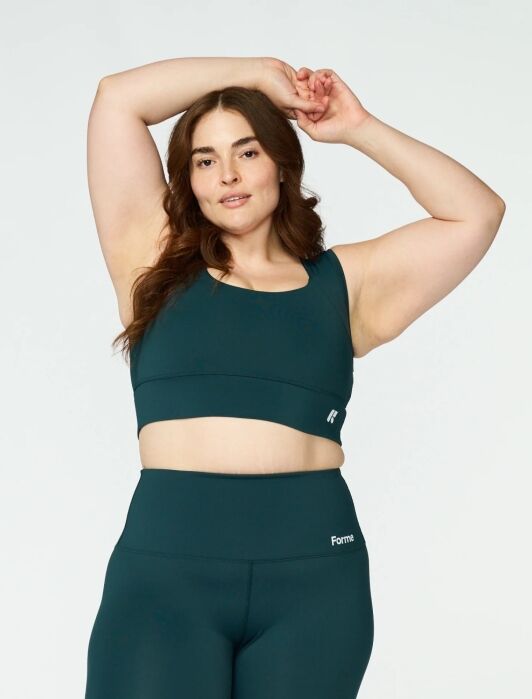 Woman in dark teal sports bra and leggings poses with one arm raised and the other resting on her head, standing against a plain light background.
