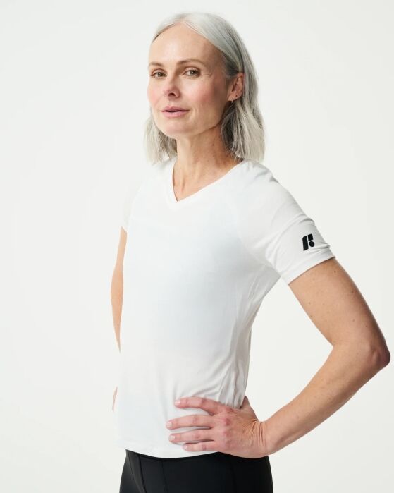 Woman with gray hair wearing a white short-sleeve shirt and black pants, standing with one hand on her hip against a plain light background.