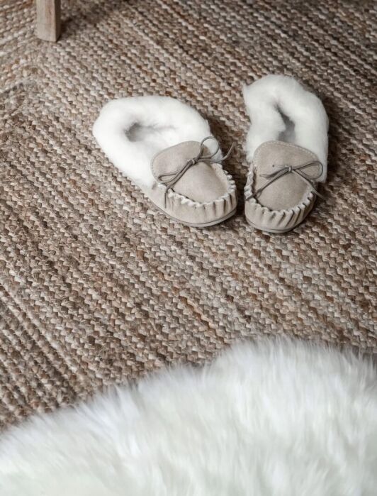 A pair of beige, fur-lined slippers rests on a textured woven rug next to the edge of a white furry mat.