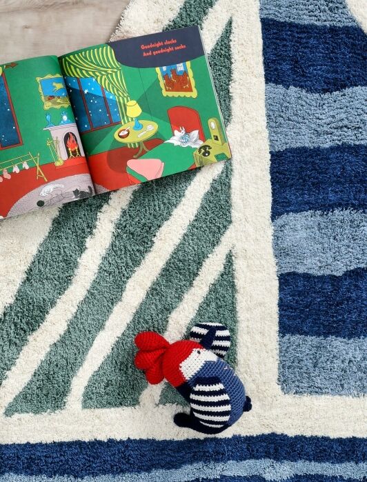 A children’s book is open on a blue and white triangular rug, next to a knitted mouse toy wearing a red hat and striped sweater.