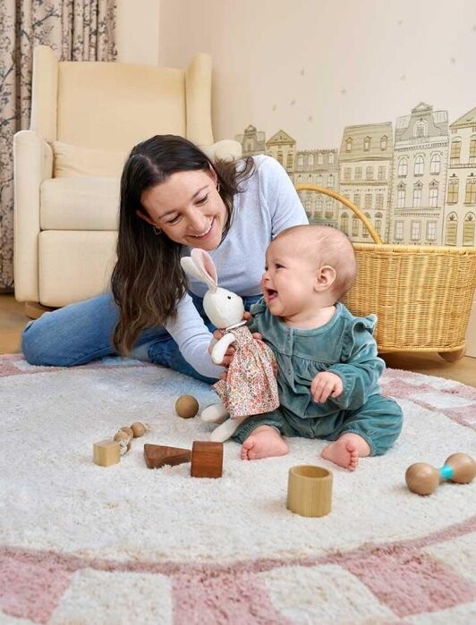An adult and a baby sit on a circular rug in a nursery, playing with a stuffed rabbit and wooden toys. A rocking sheep and a basket are in the background.