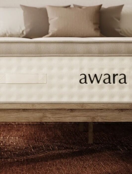 A bed with an Awara mattress on a wooden frame in a modern bedroom with a rug, pillows, and a nightstand holding a vase with flowers.