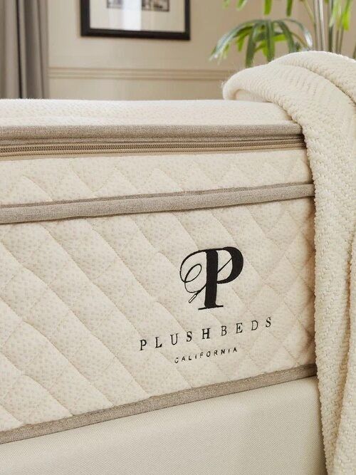 A close-up of a cream-colored PlushBeds California mattress with a textured blanket draped over the corner; a plant and part of a framed photo are visible in the background.