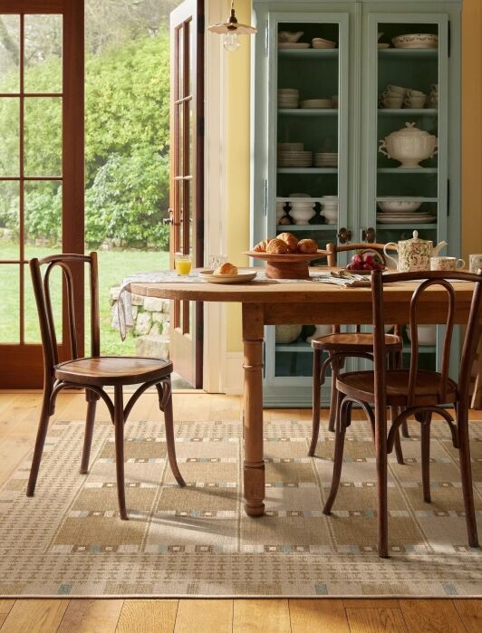 A wooden dining table with chairs is set for breakfast with croissants, fruit, and drinks. A blue cabinet with dishes and large windows are in the background.