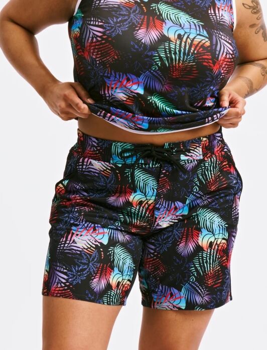 Person wearing a matching tank top and shorts set with a colorful leaf pattern on a black background, lifting the shirt slightly to show the shorts.