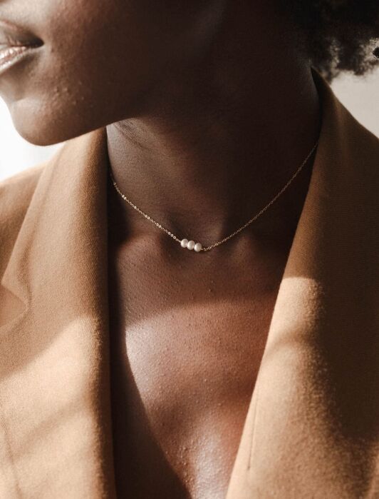 A person wearing a tan blazer and a delicate gold necklace with three small white pearls.