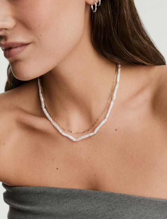 A woman wearing a strapless gray top, small hoop earring, and two layered necklaces—one with irregular white beads and one thin gold chain.