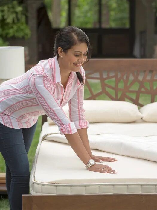 A woman presses down on a mattress with her hand while standing beside a bed frame outdoors, checking its firmness.