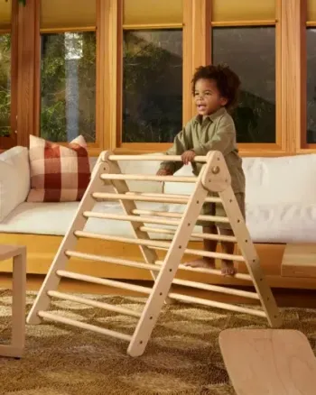 A young child stands and plays on a wooden climbing triangle in a sunlit living room with wooden accents and a white sofa.