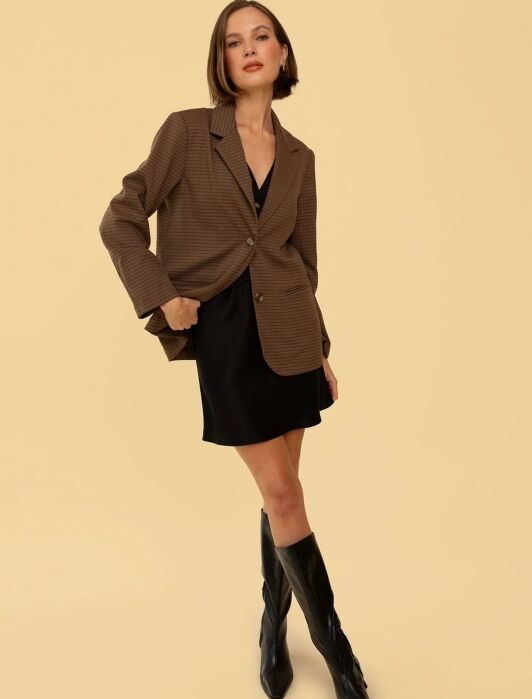 A woman stands against a beige background wearing a brown checked blazer over a black dress and knee-high black boots.