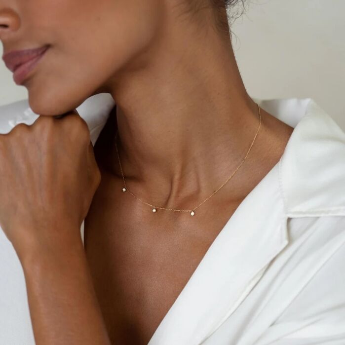 A woman in a white collared shirt wears a delicate gold necklace with three small beads, with her hand resting near her chin.