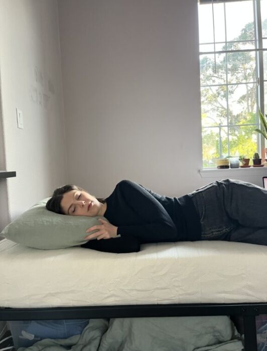 Person lying on their side on a bed with a green pillow, eyes closed, wearing a black top and gray jeans, next to a window with sunlight and potted plants visible outside.