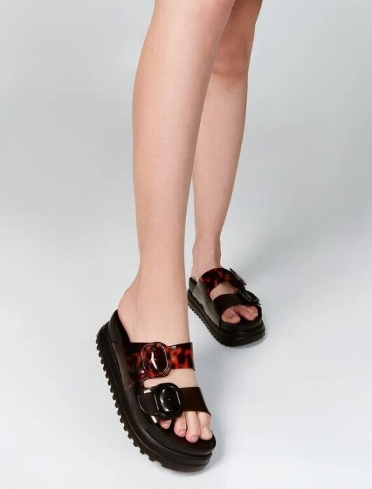 Person wearing black platform sandals with two wide, brown tortoiseshell-patterned straps and silver buckles, standing against a plain light background.