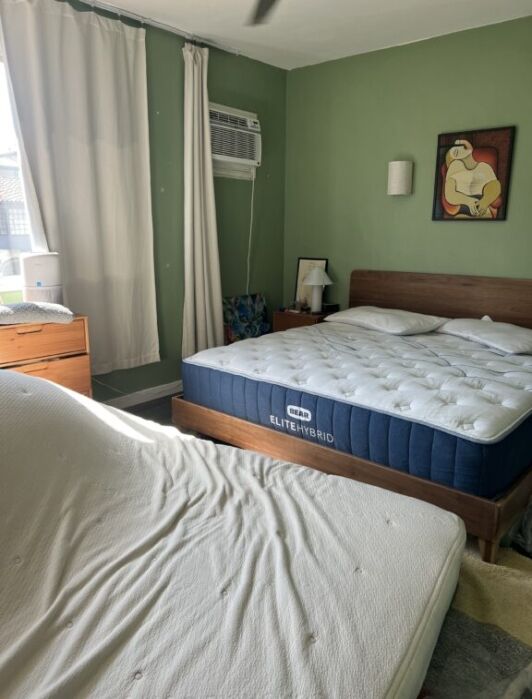 A bedroom with two beds, one upright and one at an angle, green walls, a window with a curtain, an air conditioner, a nightstand, and a colorful painting on the wall.