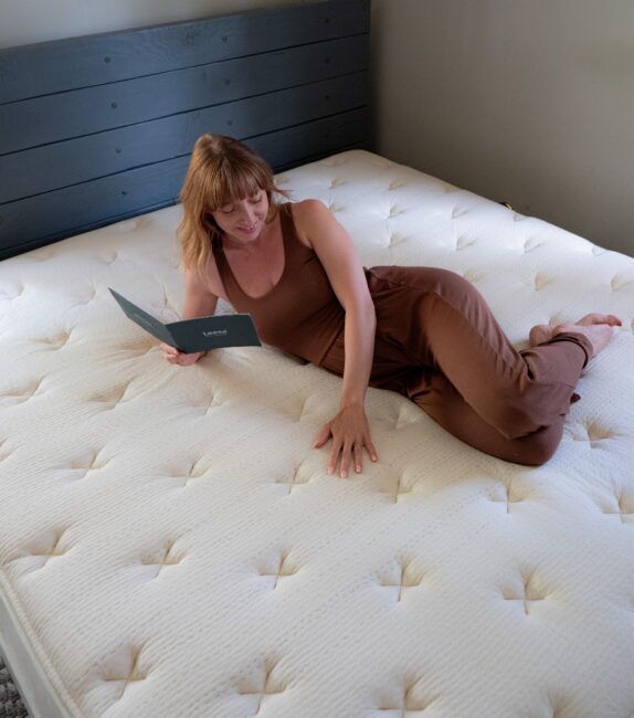 A woman in a brown outfit lies on a mattress, reading a booklet.