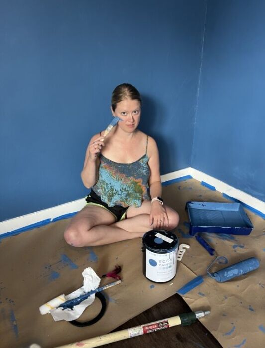 A woman sits cross-legged on the floor of a blue-painted room, holding a paintbrush with painting supplies and a paint can around her.