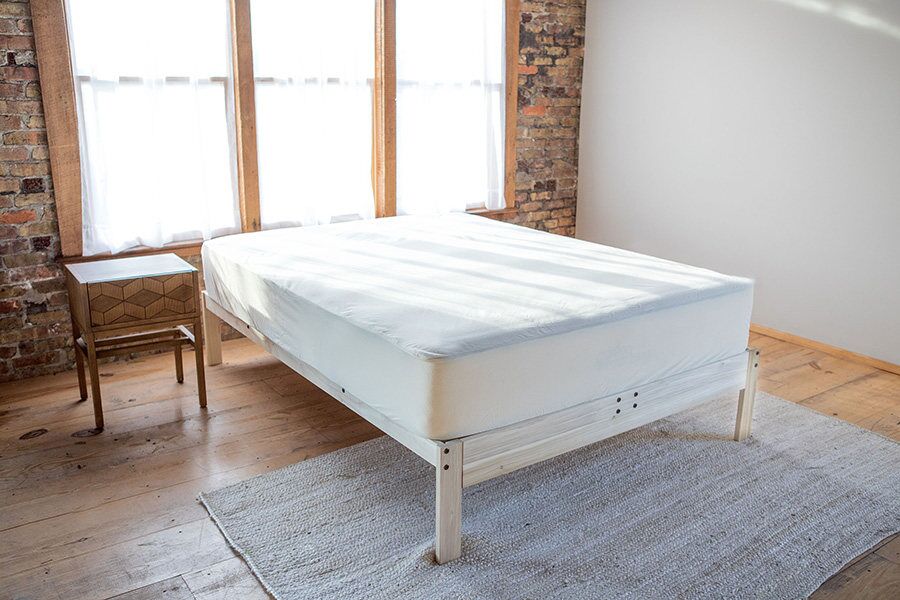 A minimalist bedroom with a bed on a light wooden frame, a small wooden nightstand, and a rug on a wooden floor, with sunlight streaming through two windows.