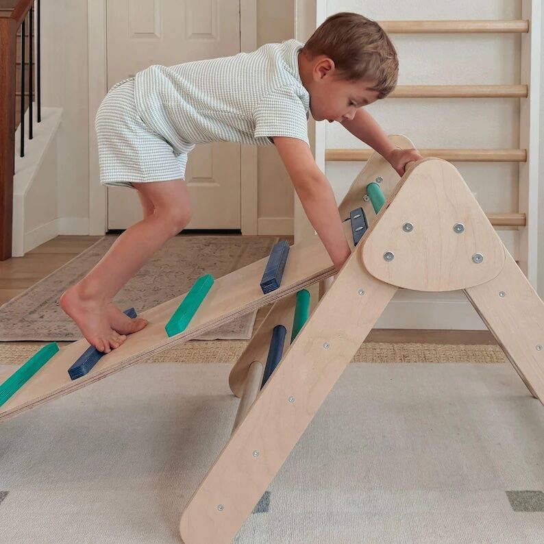 A young boy in a striped outfit climbs a wooden indoor play structure with colored climbing holds in a home setting.