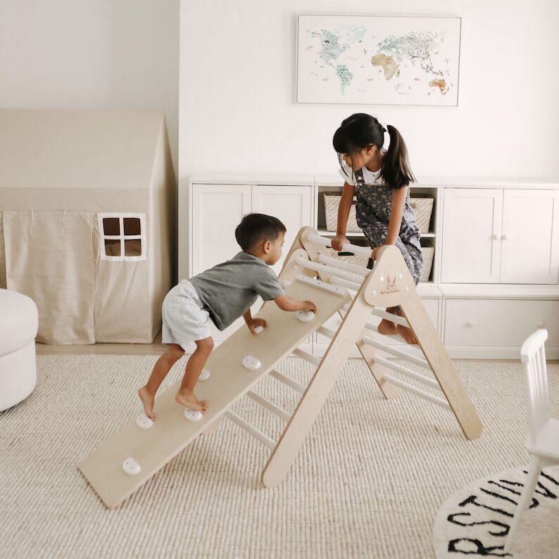 Two children play on a wooden climbing triangle in a bright, tidy room with a play tent and world map on the wall.