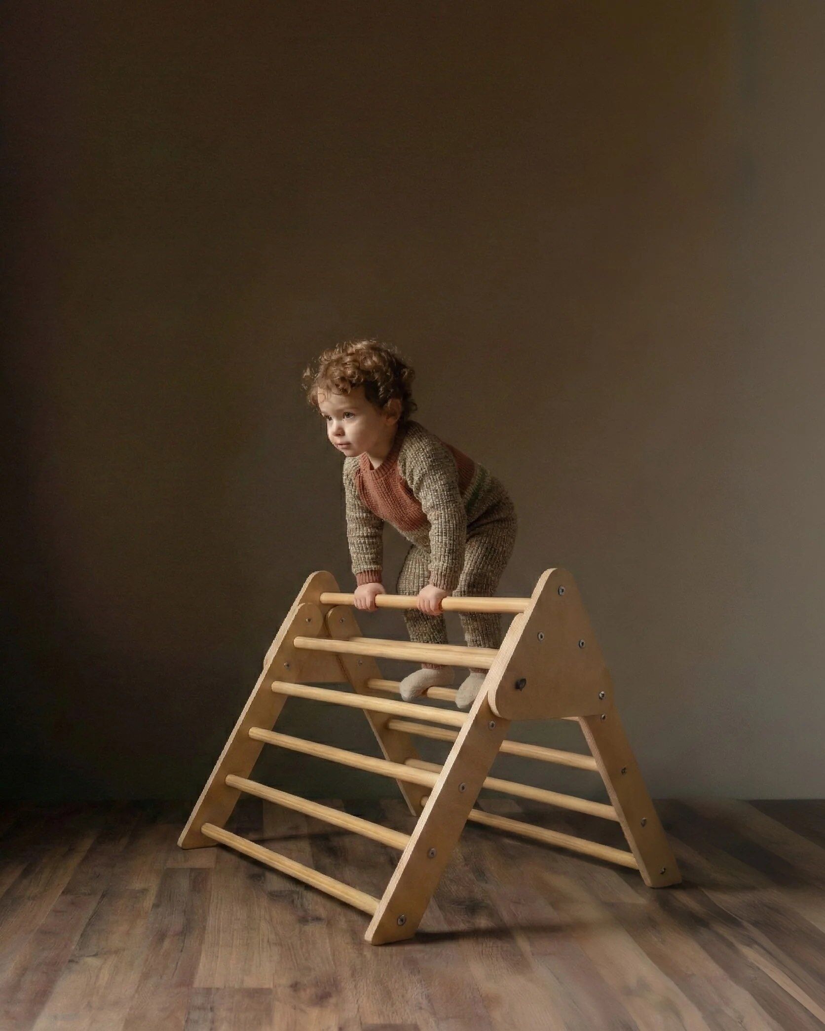 A toddler in a knitted outfit climbs on a wooden climbing structure indoors against a plain, brown background.