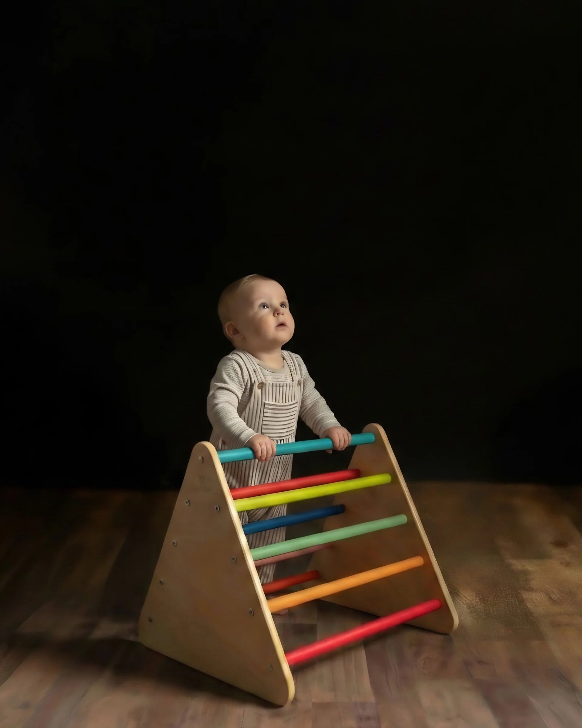 A baby stands holding a wooden climbing triangle with colorful bars, looking upward against a dark background.