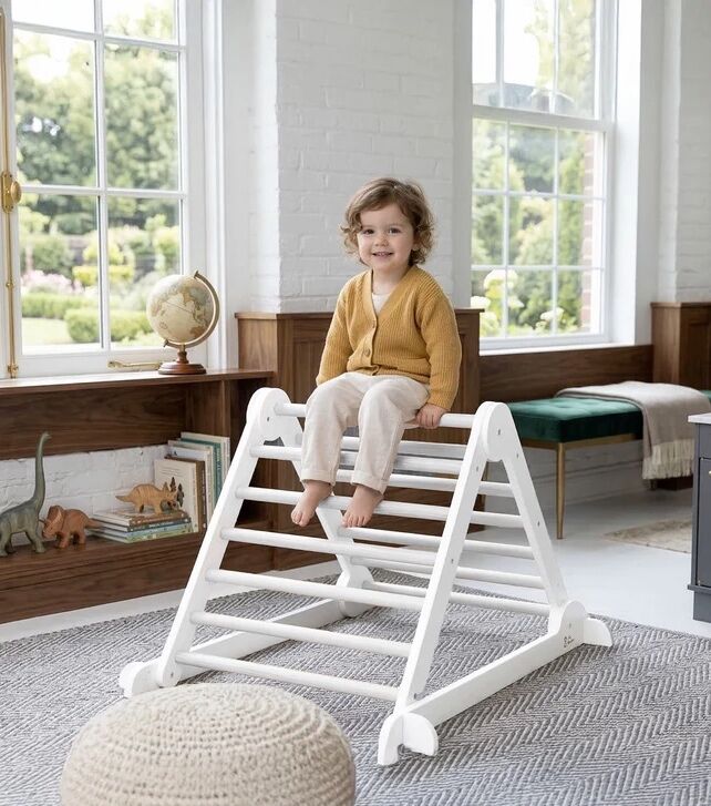 A young child sits on top of a white wooden climbing structure in a bright room with large windows and dinosaur toys on a shelf.
