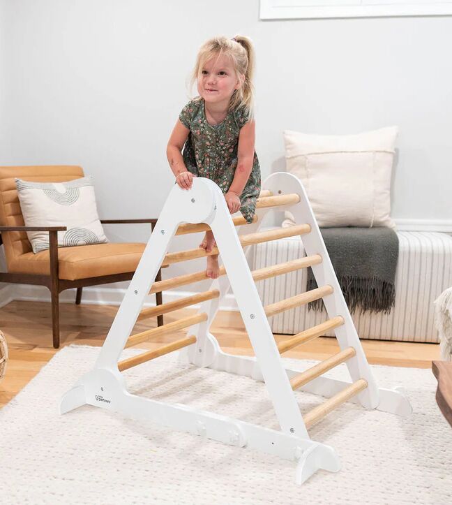 Young child in a floral dress climbs on a white wooden indoor climbing triangle in a living room with neutral decor.