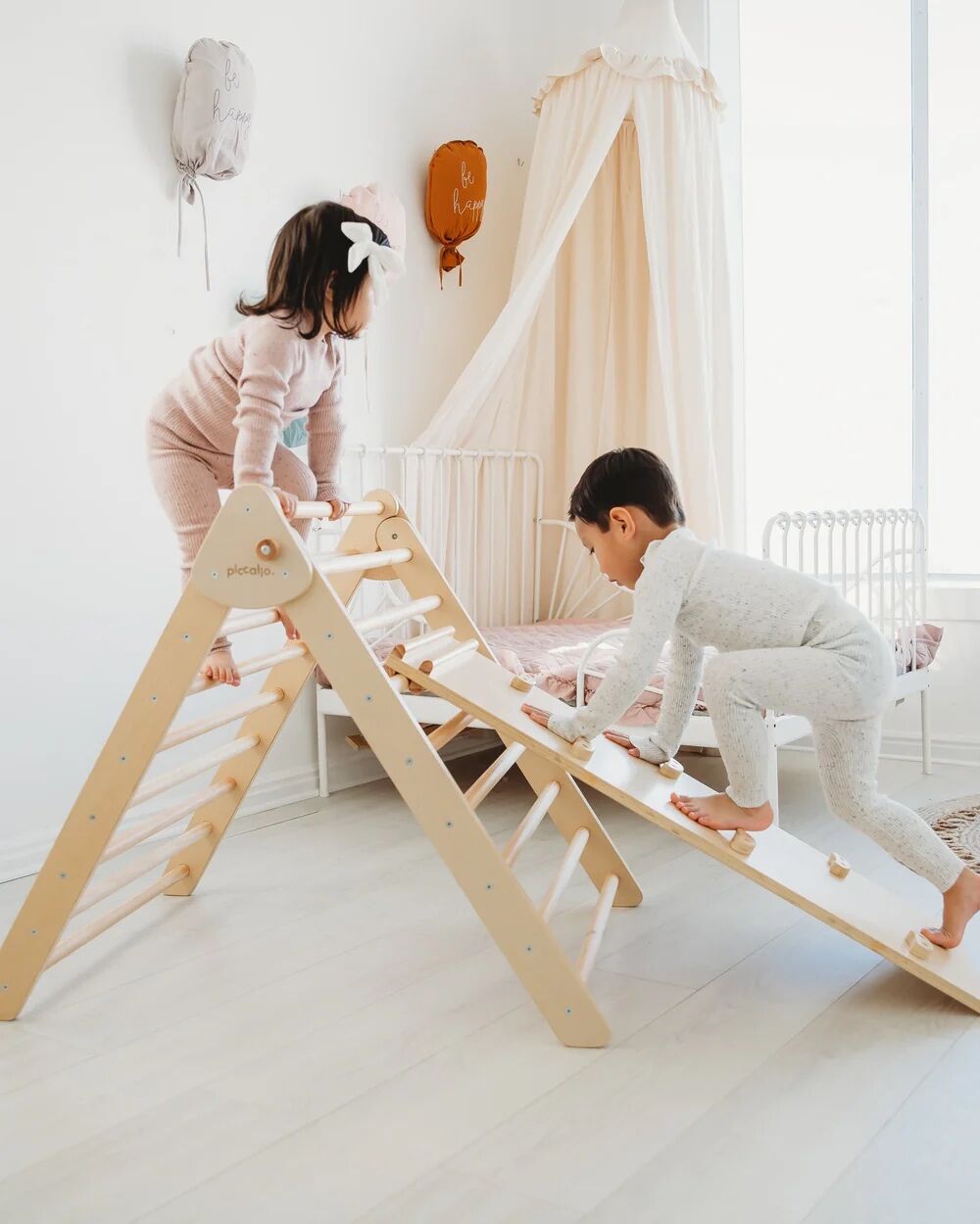 Two children in pajamas play on a wooden climbing triangle and ramp in a light-colored, softly decorated kids’ room with a small bed and canopy.