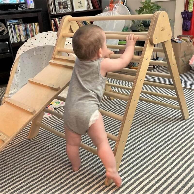A baby in a gray onesie climbs a wooden indoor triangle ladder on a striped rug, with shelves and various household items in the background.