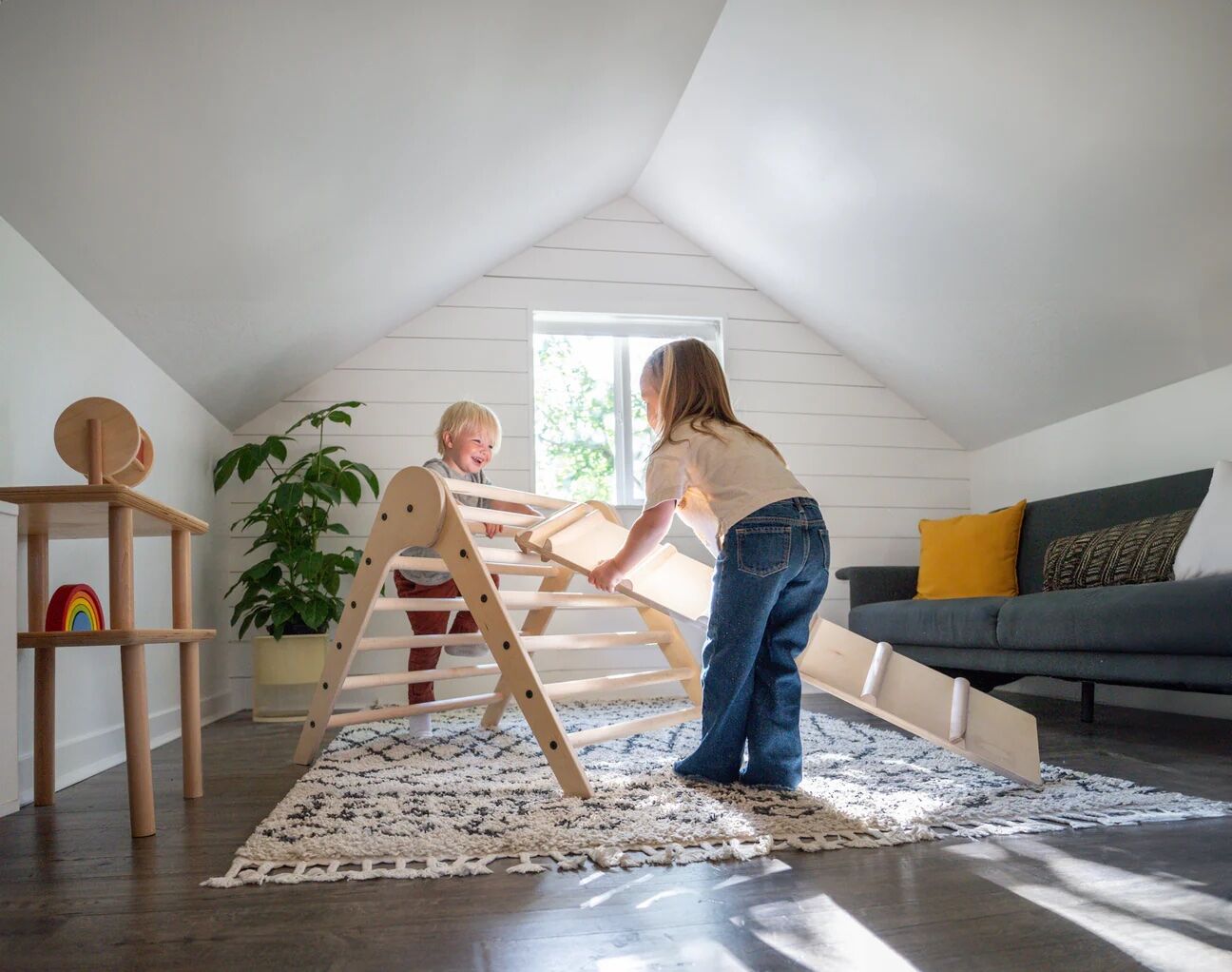 Two young children play on a wooden climbing structure in a bright, attic room with a rug, sofa, plants, and natural light from a window.