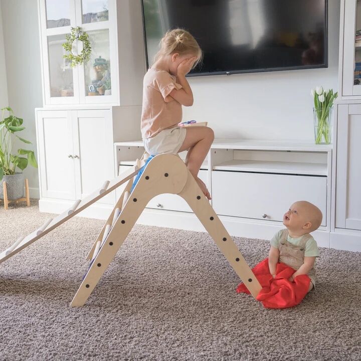 A young girl sits atop a wooden climbing structure covering her eyes, while a baby sits on the floor nearby looking up at her.