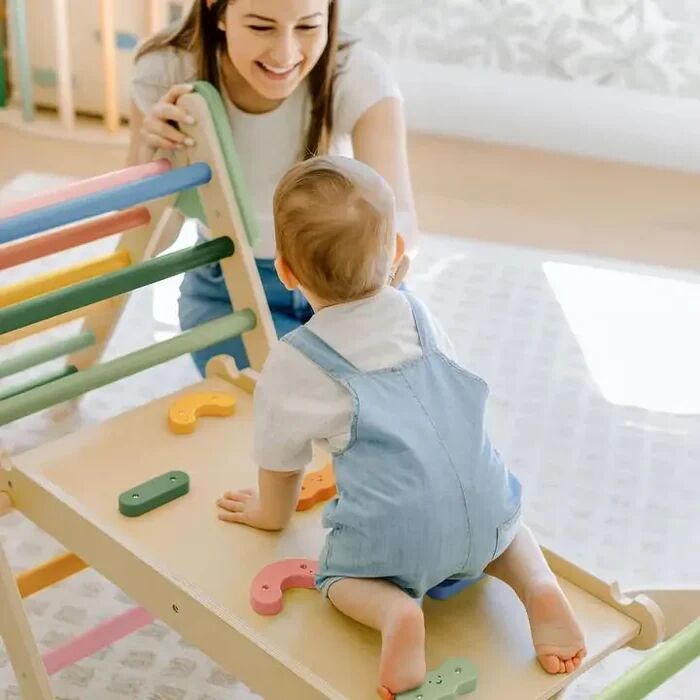 A baby in blue overalls climbs a wooden play ramp while an adult woman smiles nearby in a bright room.