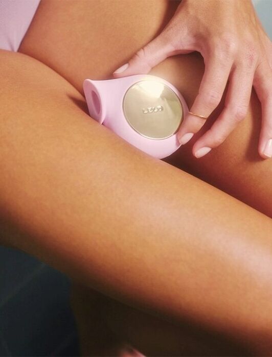 A person holding a pink and gold handheld device near their upper inner thigh while sitting.