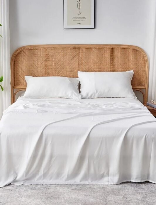 A neatly made bed with white sheets and pillows, a rattan headboard, nightstands, a plant, a lamp, and a framed artwork on the wall in a bright, modern bedroom.