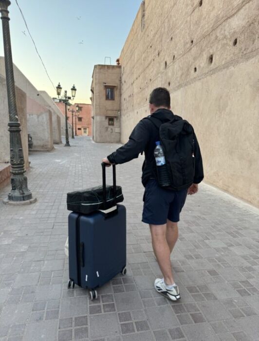 A man wearing a backpack and shorts pulls a blue suitcase down a narrow, empty street lined with beige walls and vintage street lamps.