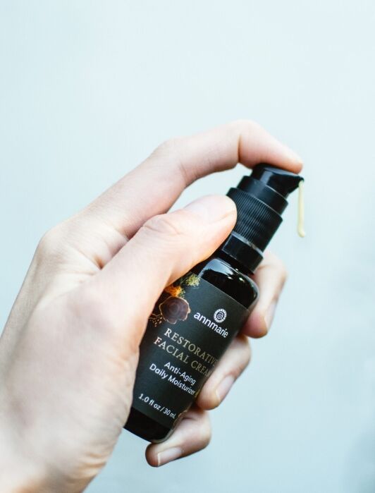 A hand holding a bottle of Annmarie Restorative Facial Cream, with a drop of cream coming out of the pump dispenser.