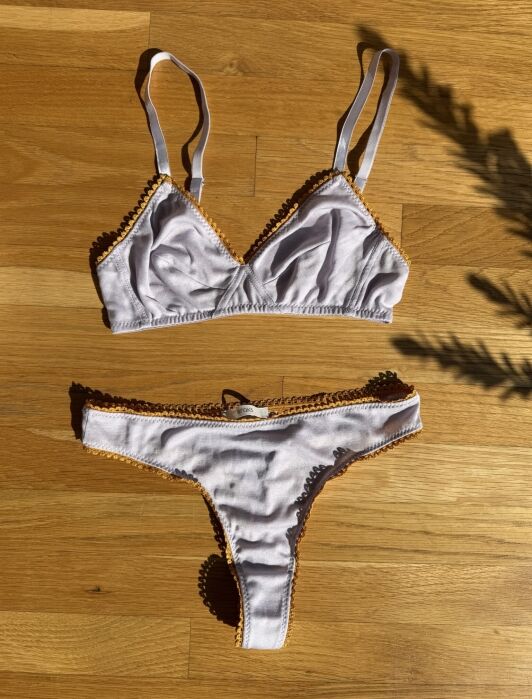 A lilac bralette and matching thong with orange trim are laid flat on a wooden floor in direct sunlight.