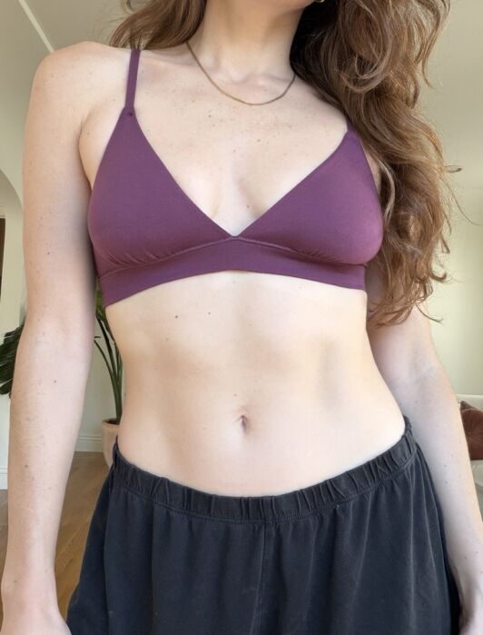 A woman wearing a purple bralette and black pants stands indoors; her face is partially out of frame.