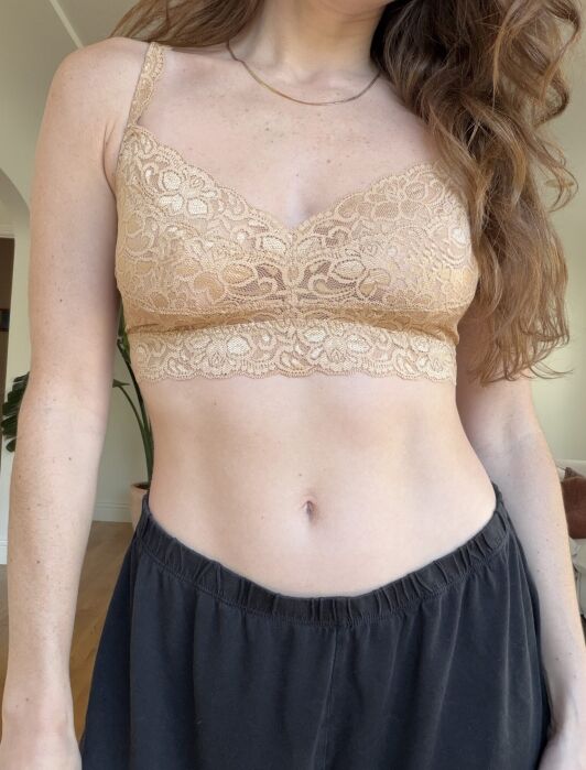 A person wearing a beige lace bralette and black pants, standing indoors with long hair partially visible.
