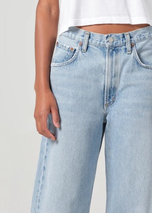 Person wearing a white cropped top and high-waisted light blue jeans, standing with one arm relaxed by their side against a plain background.
