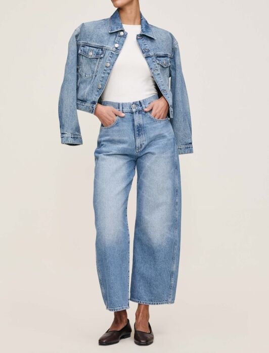 A person wearing a light blue denim jacket, white top, wide-leg blue jeans, and black shoes stands against a plain background.