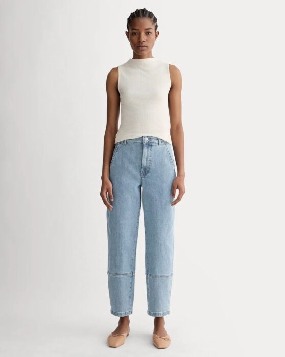 Person standing against a plain background, wearing a sleeveless beige top, light blue high-waisted jeans with seam detail, and beige flats.